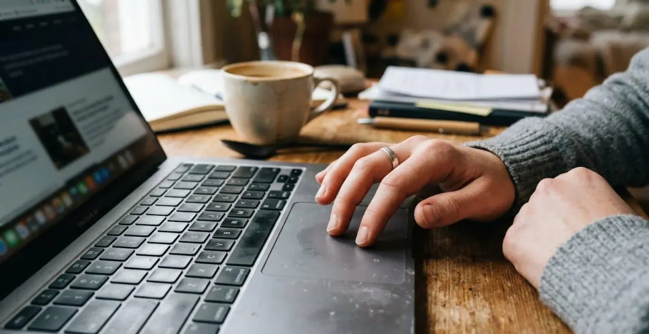 Hands on a laptop trackpad with a coffee cup nearby in a casual workspace setting