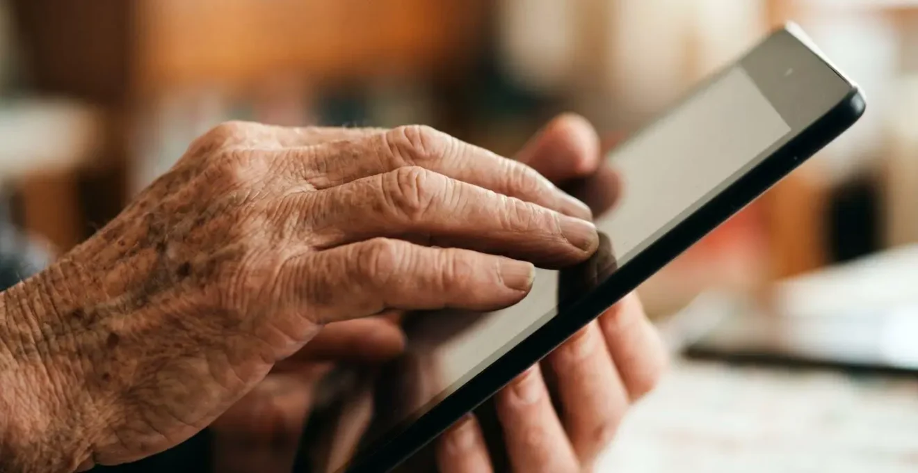 Close-up showing the weathered hands of an older person touching a tablet screen, with younger hands in the background, symbolising collaboration across generations in a UK business.